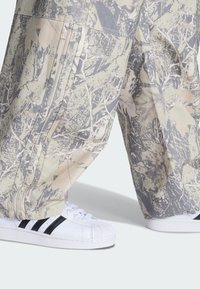Løse bukser med camouflageprint i dæmpede jordfarver med lodrette syninger; kombineret med hvide sneakers med sorte striber.