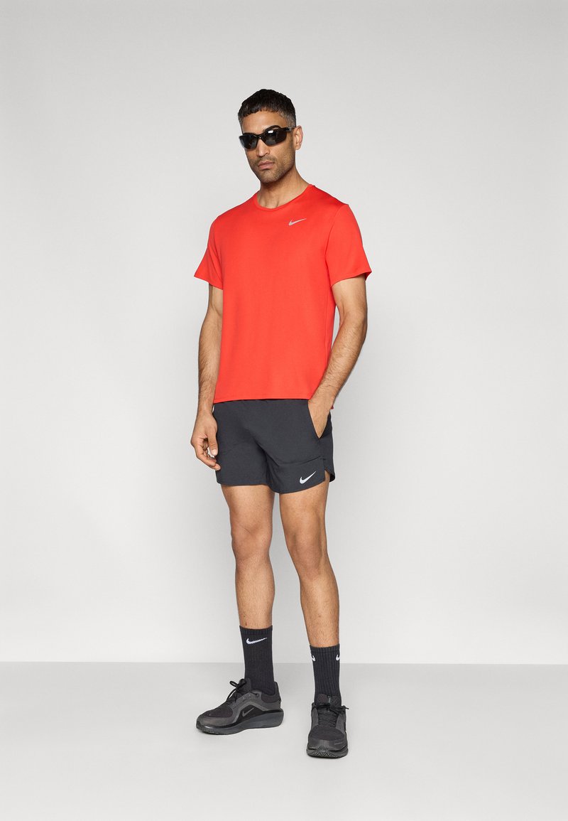 Rotes Nike-T-Shirt; schwarze Nike-Shorts; schwarze Sportschuhe; schwarze Socken; Sonnenbrille; einfarbiger Hintergrund. Fokus auf Sportbekleidung und Accessoires.