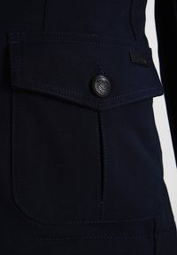 Giacca blu navy con tessuto testurizzato, grande tasca frontale con bottone e dettagli di cucitura a contrasto.