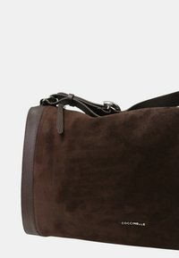 Ciemnobrązowa zamszowa torba na ramię z skórzanym wykończeniem, regulowanym paskiem, srebrną klamrą oraz małym srebrnym logo "COCCINELLE".