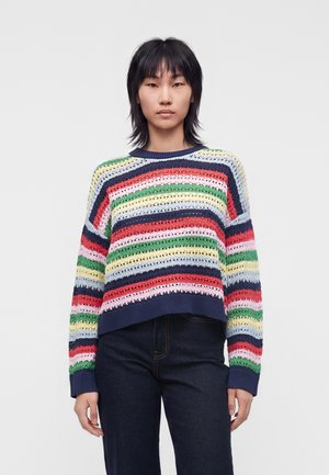 Personne debout, portant un pull ample à rayures multicolores et un jean foncé, sur un fond clair uni, faisant face avec une expression neutre.