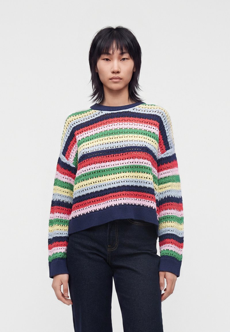 Personne debout, portant un pull ample à rayures multicolores et un jean foncé, sur un fond clair uni, faisant face avec une expression neutre.