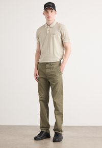 Beige polo shirt with gray accents, black cap with logo, green pants, and black sneakers. Positioned against a neutral background.