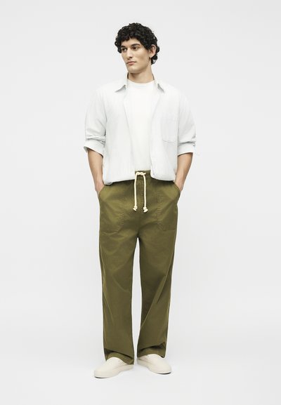 Νέος άνδρας με σγουρά μαλλιά που φοράει λευκό πουκάμισο πάνω από λευκό μπλουζάκι, παντελόνι χακί με κορδόνι και λευκά παπούτσια τύπου slip-on, τα χέρια στις τσέπες.