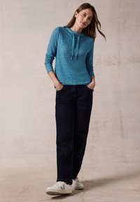 Türkiser Pullover mit Kapuze und Kordelzügen, kombiniert mit dunklen Jeans und weißen Sneakers. Glatte Textur, lässige Passform.