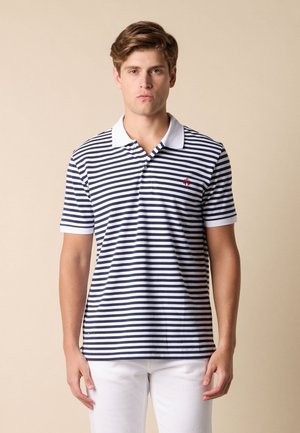 Giovane uomo che indossa una polo a righe orizzontali bianche e blu navy con colletto bianco e pantaloni bianchi, in piedi davanti a uno sfondo beige.