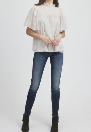 Femme portant un chemisier blanc transparent à col froufrou avec des manches papillon, un jean skinny bleu foncé et des bottines noires, debout devant un fond uni.