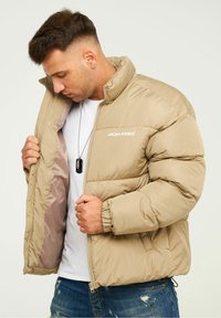 Beige Daunenjacke mit hohem Kragen, elastischen Bündchen und Reißverschluss. Mit Logo auf der Brust und glatter Innenseite. Lässiges Design.