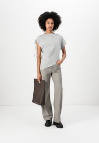 Top corto grigio a maniche corte con lacci laterali, abbinato a pantaloni a gamba larga grigi chiari. La modella tiene in mano una borsa tote marrone e indossa scarpe con plateau nere.