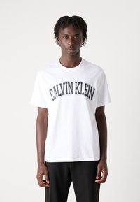 Hvit bomulls t-skjorte med "CALVIN KLEIN" trykt med fete svarte bokstaver over brystet. Standard passform med korte ermer og rund hals.