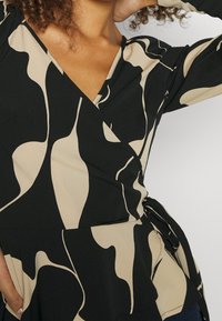 Top avvolgente nero e beige con scollatura a V profonda, maniche lunghe e motivo astratto. Include un dettaglio con fiocco laterale per la chiusura.