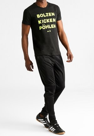 Camiseta negra con texto amarillo "BOLZEN KICKEN PÖHLEN", combinada con pantalones deportivos negros y zapatillas negras con rayas blancas.