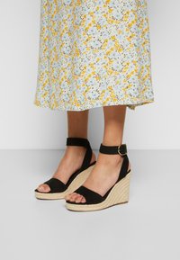 Sandalias de cuña negras con correas en el tobillo. Material de ante con base de paja trenzada. Diseño de punta abierta y acento de hebilla.
