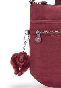 Burgundy shoulder bag with fabric texture, two zippered pockets, and a plush monkey keychain attached for added detail.