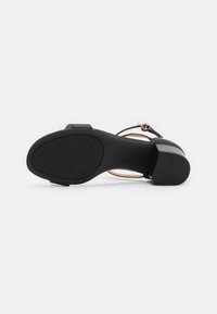 Sandale à talons hauts noire avec une semelle en caoutchouc texturé, des fines lanières et un détail de bouclerie métallique. Matière supérieure lisse avec des accents beiges.