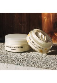 Pot de beurre corporel circulaire blanc avec un couvercle transparent, texture crémeuse visible à l'intérieur, étiqueté "SUSANNE KAUFMANN Beurre Corporel," conçu avec des accents jaunes.