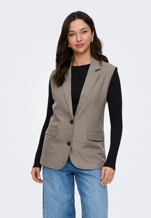 Ung kvinde med mørkt hår iført guldringe, en beige ærmeløs blazer, sort langærmet skjorte og lyseblå jeans, stående mod en hvid baggrund.
