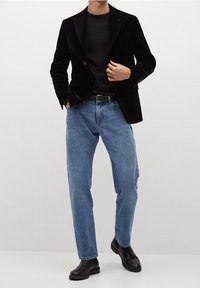 Blazer in velluto nero su una camicia nera aderente, abbinato a jeans azzurri chiari e stivaletti neri. Linee pulite e texture contrastanti.