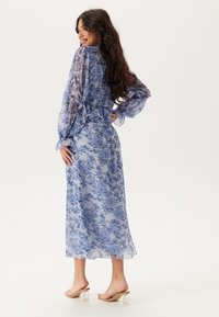 Abito maxi blu trasparente con motivo floreale, maniche lunghe a palloncino, vita arricciata e silhouette fluida. Indossato con sandali con tacco a blocco trasparente.