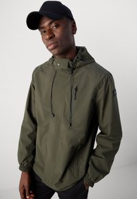 Olive green waterproof jacket with a hood, front zipper pocket, adjustable drawstrings, and elastic cuffs. Worn with a black cap.