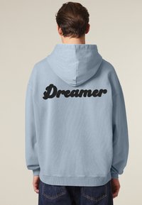 Felpa blu chiaro con una grande stampa nera "Dreamer" sul retro. Caratterizzata da una vestibilità comoda, maniche lunghe e cappuccio con cordino.