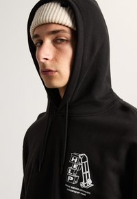 La sudadera negra presenta un gráfico en el frente con la palabra "AYUDA" y una plataforma rodante. La tela parece suave, con cordones ajustables y detalles acanalados.