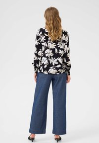 Blouse noire avec un motif floral blanc, coupe décontractée, manches longues et poignets élastiques, associée à un pantalon en denim bleu à jambes larges.