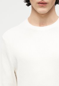 Άτομο με μεσαίου μήκους σκούρα μαλλιά που φοράει λευκό πουλόβερ με υφή πλεκτού, εμφανιζόμενο από τον λαιμό μέχρι το μέσο του στήθους.