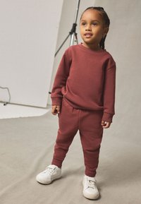 Sweat-shirt en coton rouge et joggers assortis, dotés de poignets côtelés, coupe décontractée, et baskets blanches, posés sur un fond neutre.