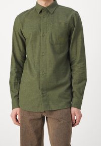 Camisa verde de manga larga con botones y un bolsillo que presenta un logo. Hecha de una tela texturizada, tiene un cuello clásico y botones cosidos.