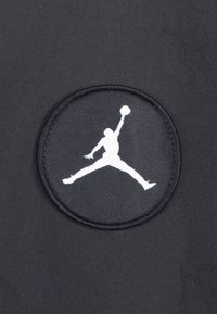 Tissu noir avec un patch circulaire arborant la silhouette blanche d'une figure sautant en tenant un ballon de basket. Design simple avec des lignes épurées.