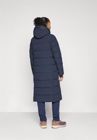 Marineblå polstret lang jakke med hette, med horisontale sømmer, elastiske ermer og en sideslisse for bedre bevegelse.