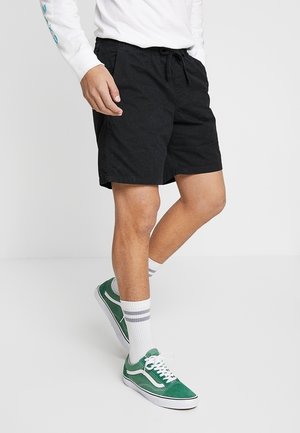 Mustat puuvillahousut, joissa on joustava vyötärö ja nauha, yhdistettyinä vihreisiin lenkkareihin sekä harmaaraidallisiin valkoisiin sukkiin.