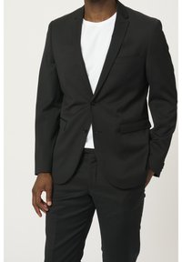 Completo black con una vestibilità slim, caratterizzato da una chiusura con un solo bottone, revers a punta e tasche laterali. Indossato sopra una t-shirt bianca.