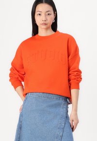 Oransje genser med preget "STUDIO"-tekst, rund hals og senkede skuldre. Kombinert med et jeansskjørt. Glatt tekstur.