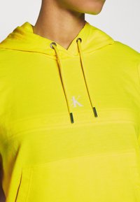 Jasnożółta bluza z kapturem i sznurkiem, z przednią kieszenią i akcentem w postaci logo. Miękka tkanina o gładkiej fakturze.