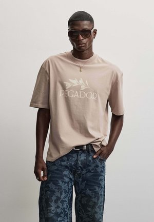 Mand med kort hår iført store solbriller, beige oversized T-shirt med bladmotiv og tekst og mørke jeans med blomsterprint.