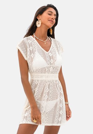 Vrouw draagt een witte gehaakte kantjurk over een bikini, geaccentueerd met grote oorbellen, een schelpenketting, een armband en een bloemenring, ogen gesloten.