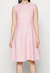 Valkoinen ilmanhihoja pinkki mekko, joka on valmistettu teksturoidusta kankaasta. Mekossa on laaja helma ja sääri hapsuilla, sekä istuva vartalonmyötäinen yläosa ja pyöreä pääntie.