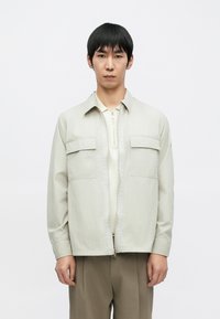 Világosszürke cipzáras dzseki két mellzsebbel, galléros nyakkal és hosszú ujjakkal. Krémszínű ing fölött viselve.