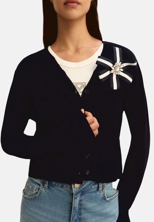 Donna che indossa un cardigan nero con bottoni con una spilla a nastro bianca e nera e una maglietta bianca sotto, abbinato a jeans blu.