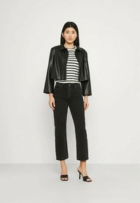 Žena v černé zkrácené kožené bundě, černých vysokých džínách, pruhované košili a černých sandálech na podpatku stojí proti jednoduché zdi.