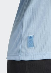Lyseblå athletisk skjorte med vertikale striper, et teksturert stoff, og en blå etikett med logo og tekst ved kanten.