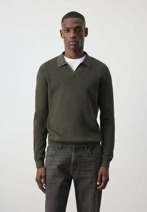 Hombre de pie, con suéter de cuello y manga larga verde oscuro sobre camisa blanca y vaqueros gris oscuro contra un fondo claro liso.