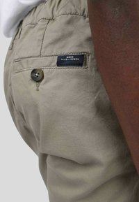 Pantalons beige clair avec une finition texturée, dotés d'une fermeture à bouton et d'une poche arrière ornée d'une étiquette de marque noir et blanc.