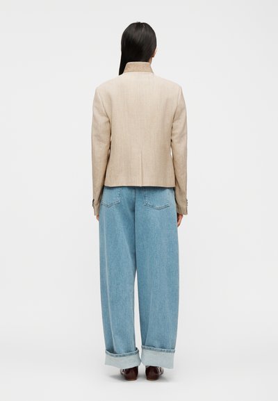 Kvinde med langt mørkt hår iført beige blazer og lyseblå bukser med vide ben, stående med ryggen til på en ensfarvet hvid baggrund.