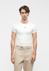 Valge lühikeste varrukatega slim-fit T-särk, millel on rinna peal väike must logo, koos beeži kõrge vöökohaga pükstega.