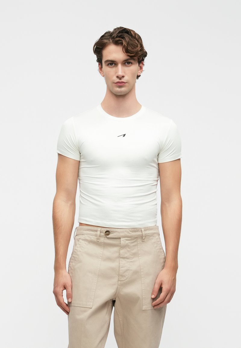 Valge lühikeste varrukatega slim-fit T-särk, millel on rinna peal väike must logo, koos beeži kõrge vöökohaga pükstega.