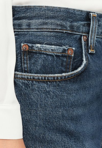 Denim jeans with a dark blue wash, featuring a small front pocket, copper rivets, and a frayed hem. Visible stitching in orange.