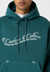 Teal hoodie met trekkoorden, voorzak en witte tekst: "Couture Culture, Elevate Your Style, Define Your Couture." Tekst gebogen en gestileerd.